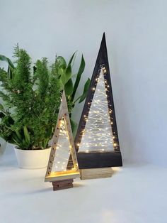 two wooden christmas trees with fairy lights on them sitting next to a potted plant