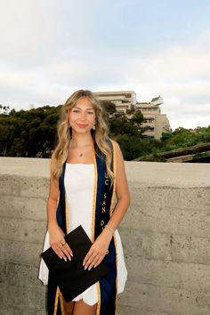 a woman standing next to a cement wall wearing a blue and gold graduation gown with her hands in her pockets