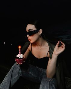 a woman sitting down holding a cupcake with a candle in her hand and wearing sunglasses