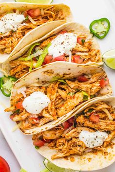 three shredded chicken tacos on a white plate with limes and tomatoes in the background