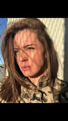 a woman with her hair blowing in the wind while wearing a camouflage jacket and looking at the camera