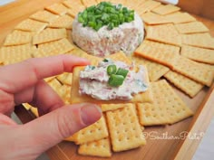 someone is holding their finger over crackers with cheese and vegetables on them, next to a bowl of dip