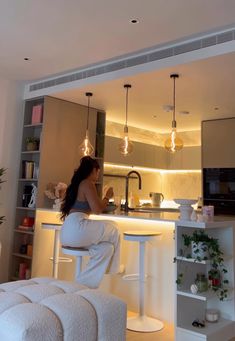 a woman sitting on a stool in front of a kitchen counter with lights hanging from the ceiling