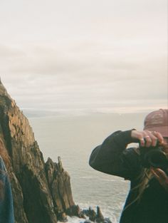 two people standing on top of a mountain next to the ocean and one person holding a camera