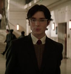 a man wearing glasses and a suit standing in a hallway with other people walking up the stairs