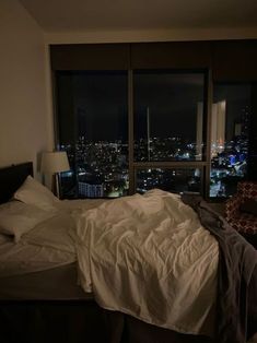 an unmade bed in front of a large window with city lights lit up at night