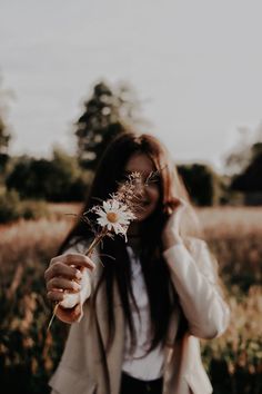 a woman standing in a field holding a flower up to her face and looking at the camera