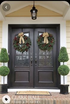 two christmas wreaths on the front door of a house with potted topiary trees