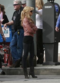 a woman in black pants and boots standing next to a man on a sidewalk with other people