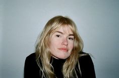 a woman with long blonde hair wearing a black shirt and looking at the camera while standing in front of a white wall