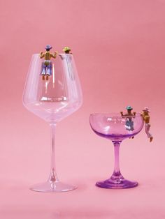 two wine glasses with figurines in them on a pink background