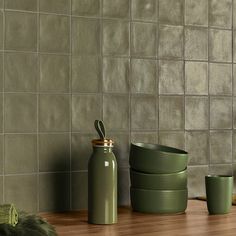 green dishes and cups are sitting on a wooden table next to a wall with grey tiles