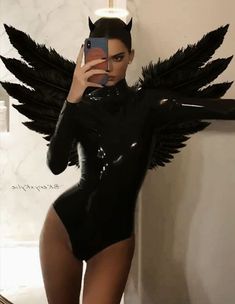 a woman in a black bodysuit taking a selfie with her cell phone while wearing angel wings