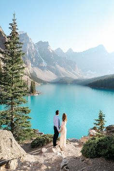 a man and woman standing on top of a mountain next to a lake