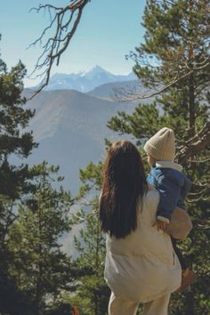 a woman holding a child in her arms while looking at the mountains and pine trees