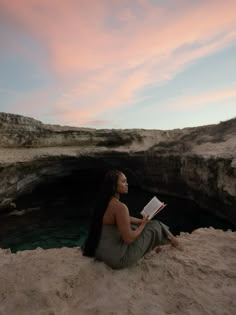 a woman sitting on top of a sandy beach next to the ocean reading a book