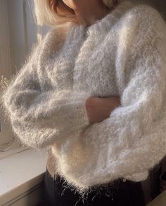 a woman standing in front of a window wearing a white sweater