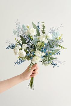 a person holding a bouquet of white and blue flowers with greenery in their hand