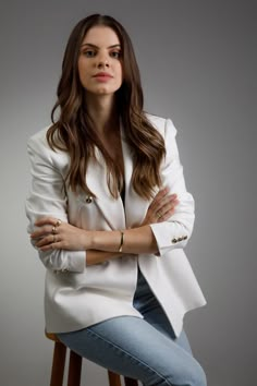 a woman sitting on top of a wooden stool wearing a white blazer and jeans