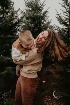 a woman holding a baby in her arms and smiling at the camera with trees behind her