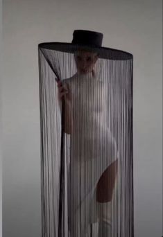 a woman in white is holding a black hat and standing behind a sheered curtain