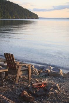 two wooden chairs sitting on top of a beach next to a fire pit and water