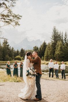 a bride and groom kissing in front of their wedding party at the river's edge