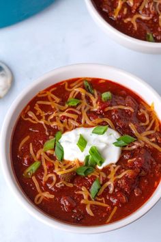 two bowls of chili with sour cream and green garnish on top, sitting next to each other