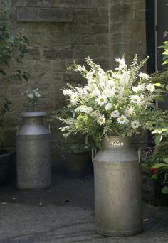 some white flowers are in large metal vases on the side of a building near potted plants