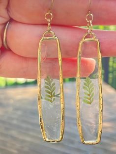 a pair of gold - plated earrings with pressed glass and green leaves is held by a hand