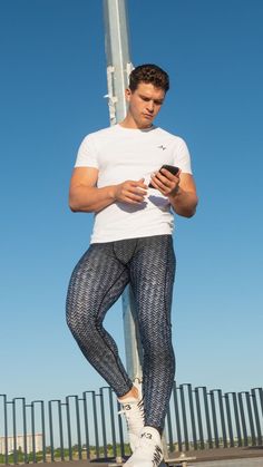 guy wearing a white t-shirt and carbon fibre print leggings and white sneakers holding and looking at his phone on a sunny day while standing up against a metal pole outside Casual Nylon Tights For Gym, Casual Nylon Leggings For Sports, Casual High Stretch Tights For Jogging, Casual Nylon Training Leggings, Casual Nylon Leggings For Training, Casual Nylon Running Leggings, Casual Compression Pants With Breathable Fabric, Casual Nylon Tights For Training, Casual Moisture-wicking Sports Tights