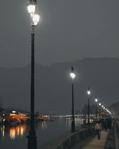 people are walking along the water at night with street lights on either side of them