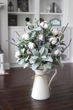 a white vase filled with flowers and greenery on top of a wooden table next to a book shelf