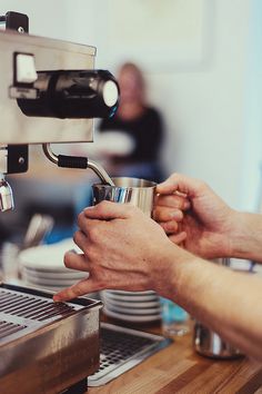 two people are holding coffee cups in front of an espresso machine while another person is sitting at the counter