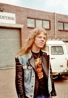 a young man with long hair standing in front of a building wearing a leather jacket