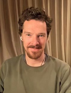 a man with curly hair and beard sitting in front of a curtained window, smiling at the camera