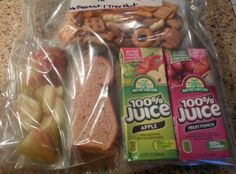 two packaged sandwiches in plastic bags on a counter top next to fruit punch and apple slices