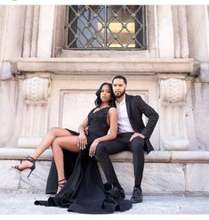 a man and woman are sitting on a bench in front of a building, posing for the camera