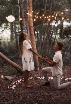 a man kneeling down next to a woman in the woods holding hands with string lights strung above them