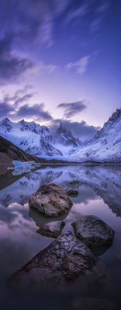 the mountains are reflected in the still water at dusk, with rocks on the shore