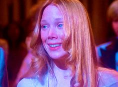 a woman with blonde hair and blue eyes smiles at the camera while standing in front of other people