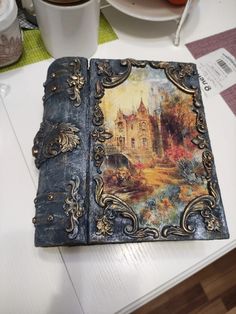 an old book with a painting on it sitting on a table next to a cup of coffee