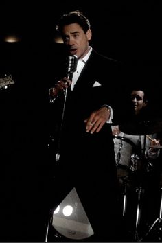 a man in a tuxedo singing into a microphone with other musicians behind him