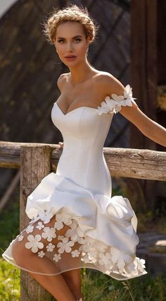 a woman in a short white dress leaning on a wooden fence with her legs crossed