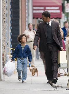 a man in a suit and tie walking with a small child on a sidewalk while people walk by