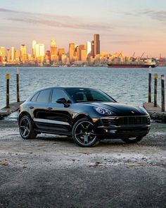 a black porsche cayen is parked in front of the water with a city skyline behind it