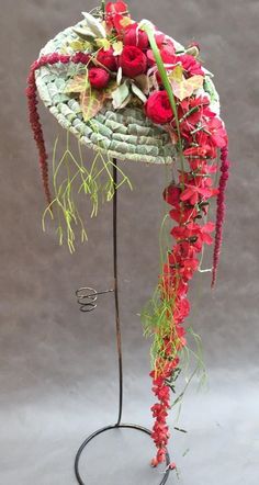 a hat with red flowers and greenery is on a metal stand in front of a gray background