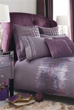 a bed with purple comforters and pillows in a room next to a lamp on a table