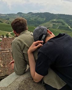 two people sitting on top of a stone wall looking at the hills and valleys in the distance