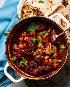 a pot filled with meat and beans next to tortilla bread on a blue towel